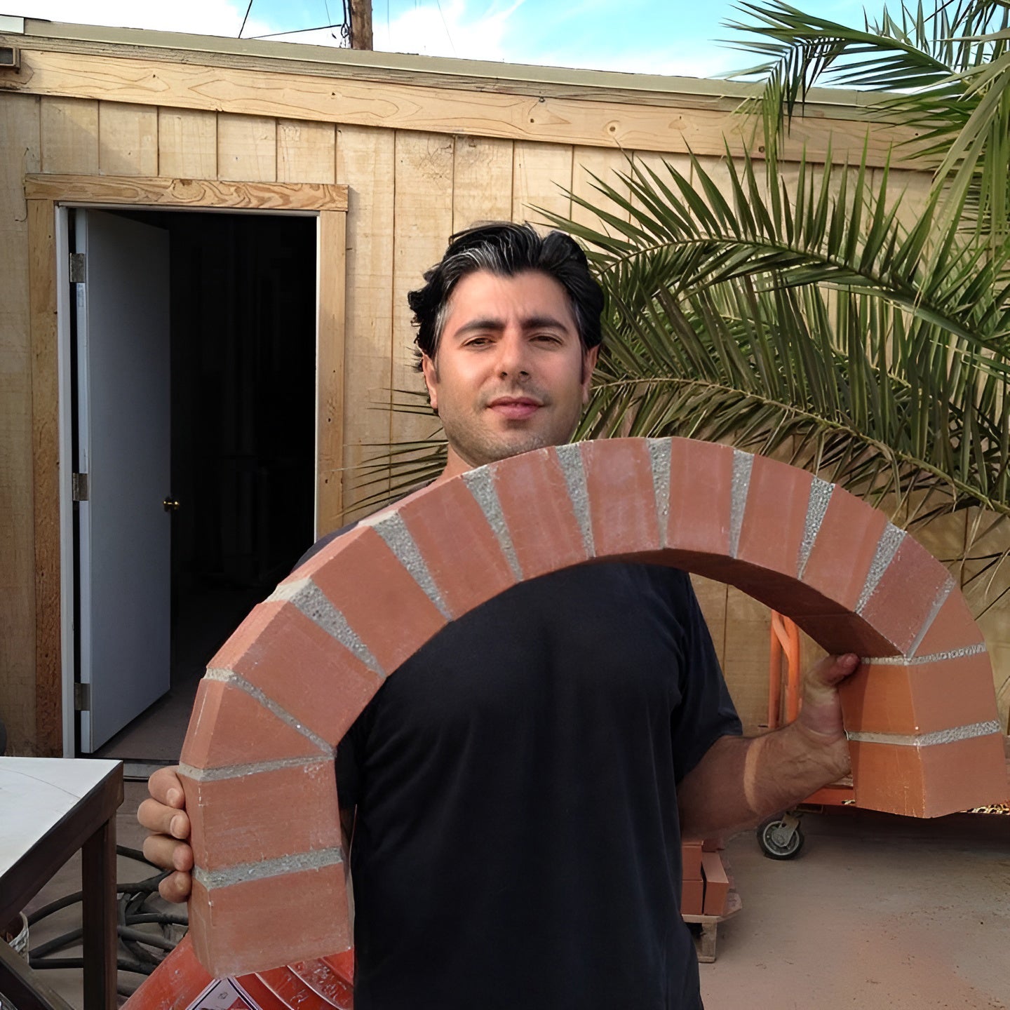 Giuseppe Crisa, Forno Classico's owner, holding a brick archway in front of a wooden shed with a palm tree in the background