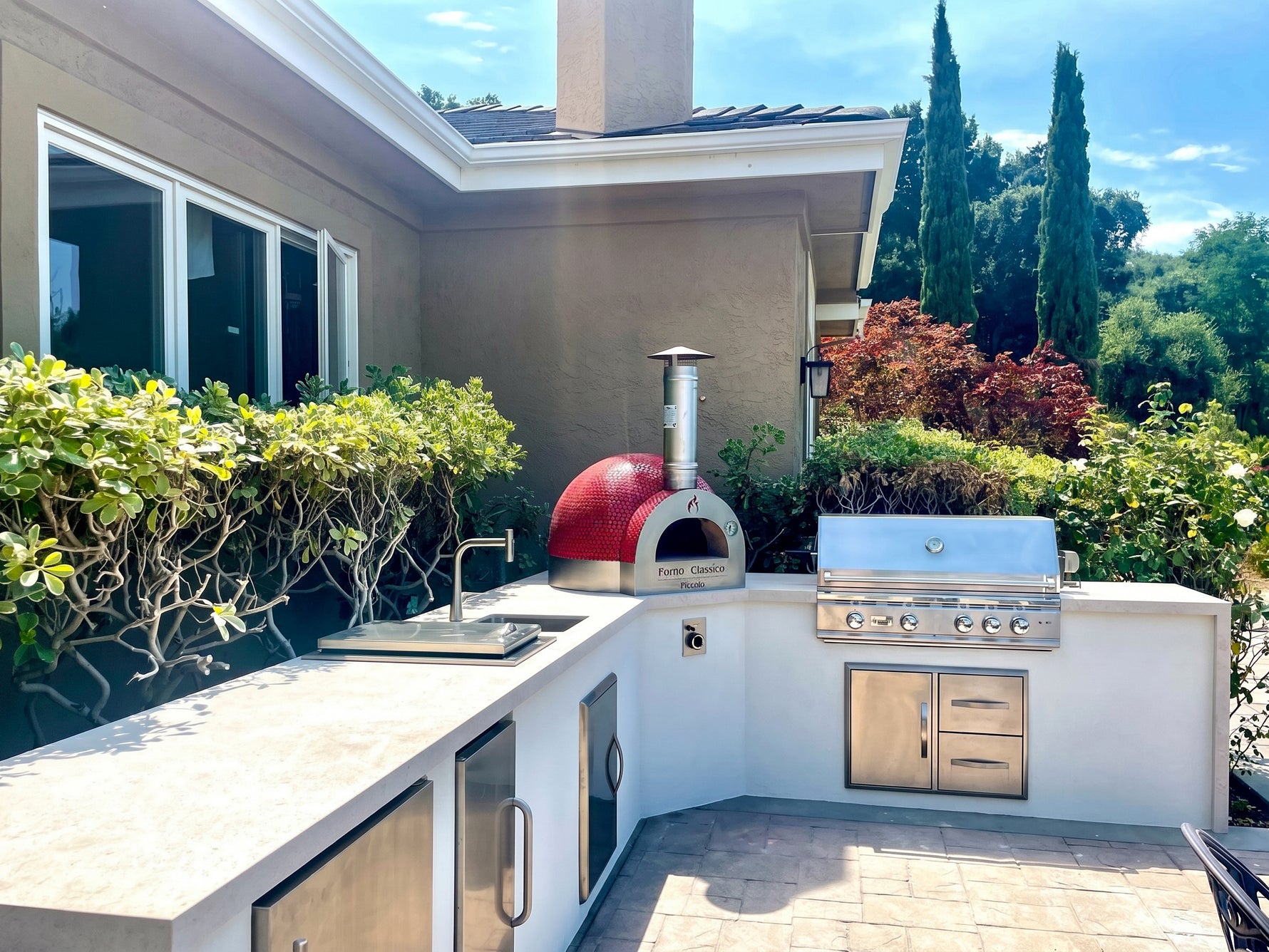 Outdoor kitchen with Forno Classico Piccolo 65 pizza oven, grill, and cabinets in a residential setting.